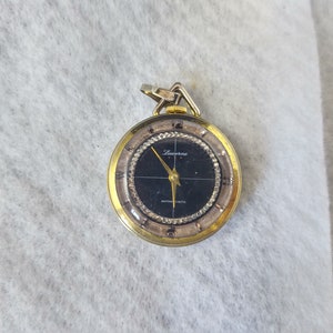 May include: A gold-toned Lucerne pocket watch with a black face and gold hands. The watch has a round face with a silver-colored bezel and a small loop at the top for attaching a chain. The watch face reads "Lucerne" and "ANTIMAGNETIC".