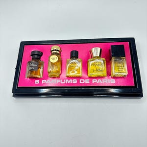 以下が含まれることがあります： ピンク色の内装の黒いディスプレイケースに入った、5つのミニチュア香水ボトルのセット。ボトルは様々な形とサイズで、金と黒のアクセントが付いています。「5 PARFUMS DE PARIS」の文字がピンク色の背景に印刷されています。