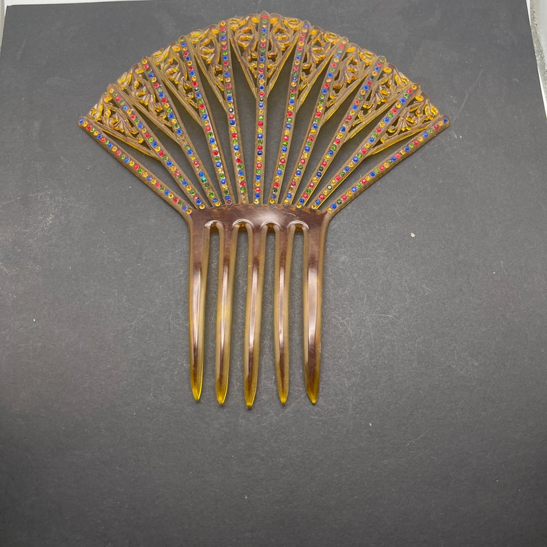 May include: A brown plastic hair comb with a decorative fan-shaped design. The design features intricate details and is accented with colorful rhinestones.