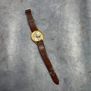 Pu&ograve; includere: Un orologio da polso in pelle marrone con cassa dorata e quadrante bianco. Il quadrante presenta un personaggio dei cartoni animati e indici orari in numeri romani neri.