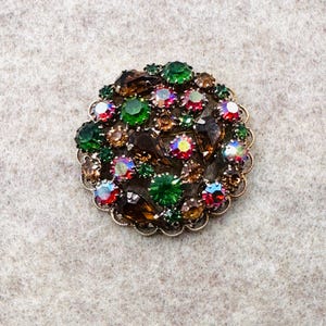 May include: A round vintage brooch with a gold-tone filigree edge. The brooch is adorned with a variety of colorful rhinestones in shades of green, brown, red, and iridescent. The stones are arranged in a clustered pattern.