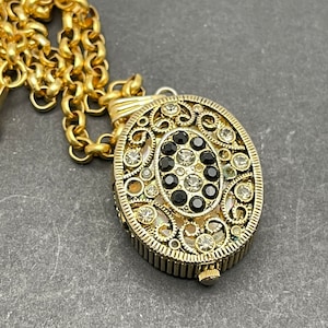 May include: Gold-toned oval pendant necklace with intricate filigree design. The pendant is adorned with clear and black crystals, and hangs from a matching gold chain. The necklace has a vintage or antique aesthetic.