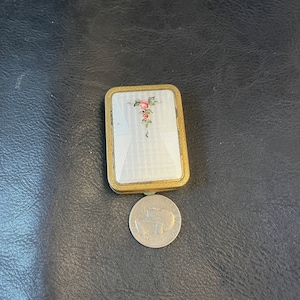 Puede incluir: Un pequeño estuche rectangular de color dorado con una tapa de esmalte blanco. La tapa tiene un diseño floral con flores rosas y verdes. El estuche es aproximadamente del tamaño de una moneda de euro.