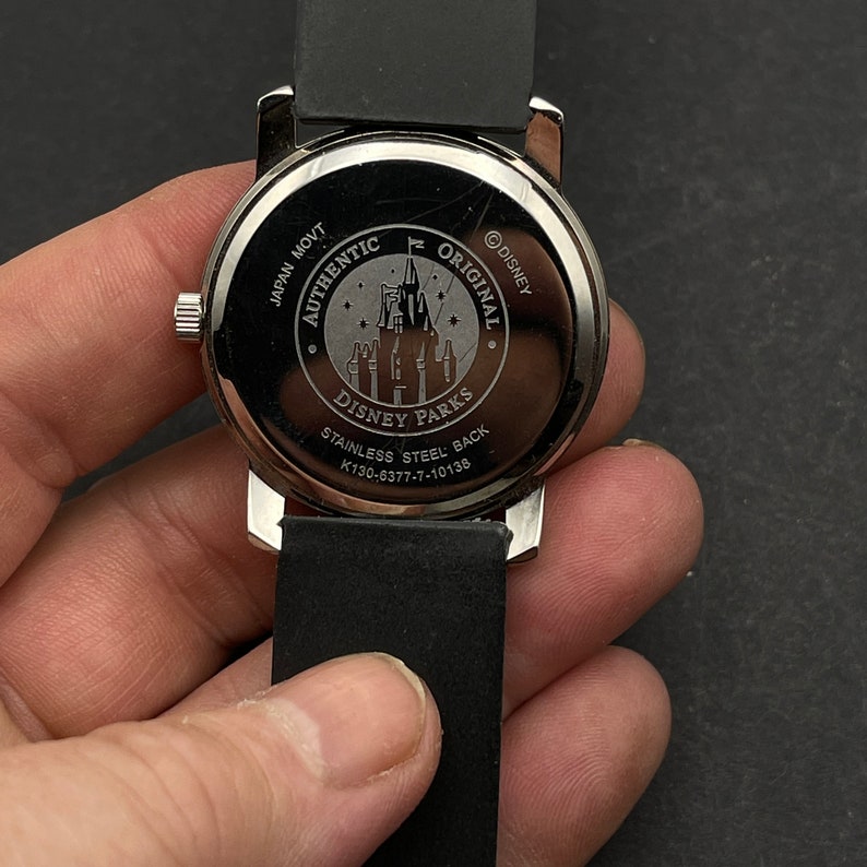 May include: The back of a silver watch with a black band. The watch face has a Disney Parks logo with the words "Authentic Original" and "Stainless Steel Back" printed on it.