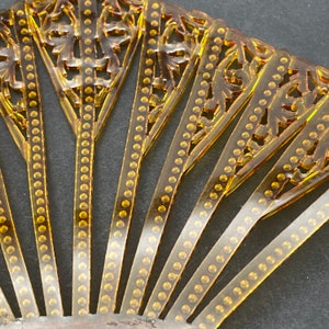 May include: A gold-colored, decorative fan made of thin, translucent plastic. The fan is made up of multiple sections, each with a delicate, floral design and a row of small, round beads.