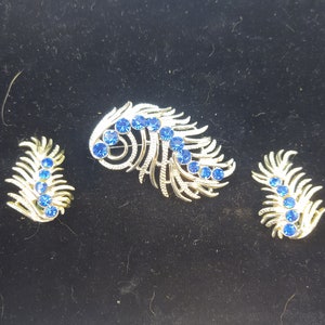 May include: A vintage jewelry set featuring a brooch and matching earrings. The set has a silver-tone metal finish and is adorned with vibrant blue rhinestones. The design resembles a stylized feather or leaf pattern, adding a touch of elegance.