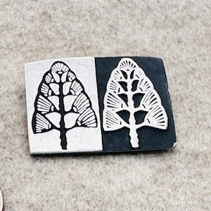 Puede incluir: Un broche rectangular de metal plateado con un diseño de árbol. El broche está dividido en dos secciones, una blanca y otra negra, cada una con un árbol estilizado con detalles intrincados. El diseño del árbol es en blanco y negro.