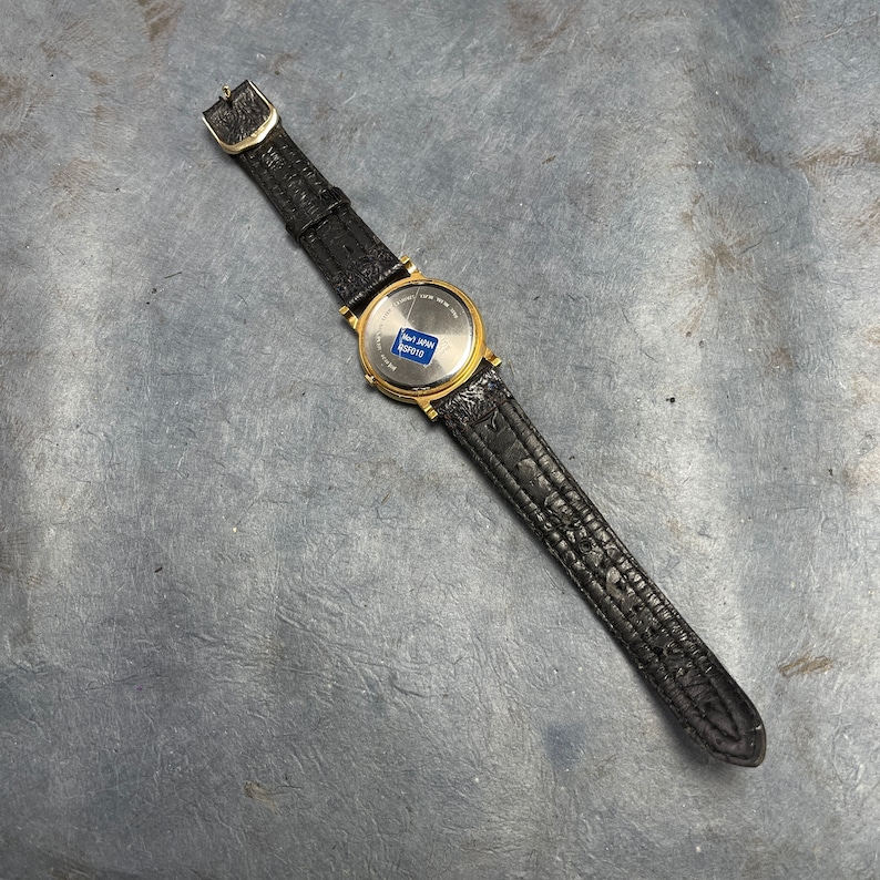 Pu&ograve; includere: Un orologio da polso dorato con un cinturino in pelle nera. Il quadrante dell'orologio &egrave; coperto da un vetro trasparente e il retro dell'orologio ha un adesivo blu che dice "Not Japan  KS2018".