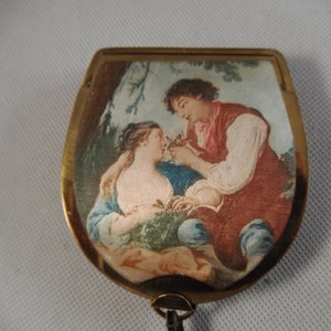 May include: A vintage compact mirror with a gold-toned frame. The mirror's surface features a romantic scene of a man and woman in a garden setting, rendered in soft colors. The compact has a small loop at the bottom.