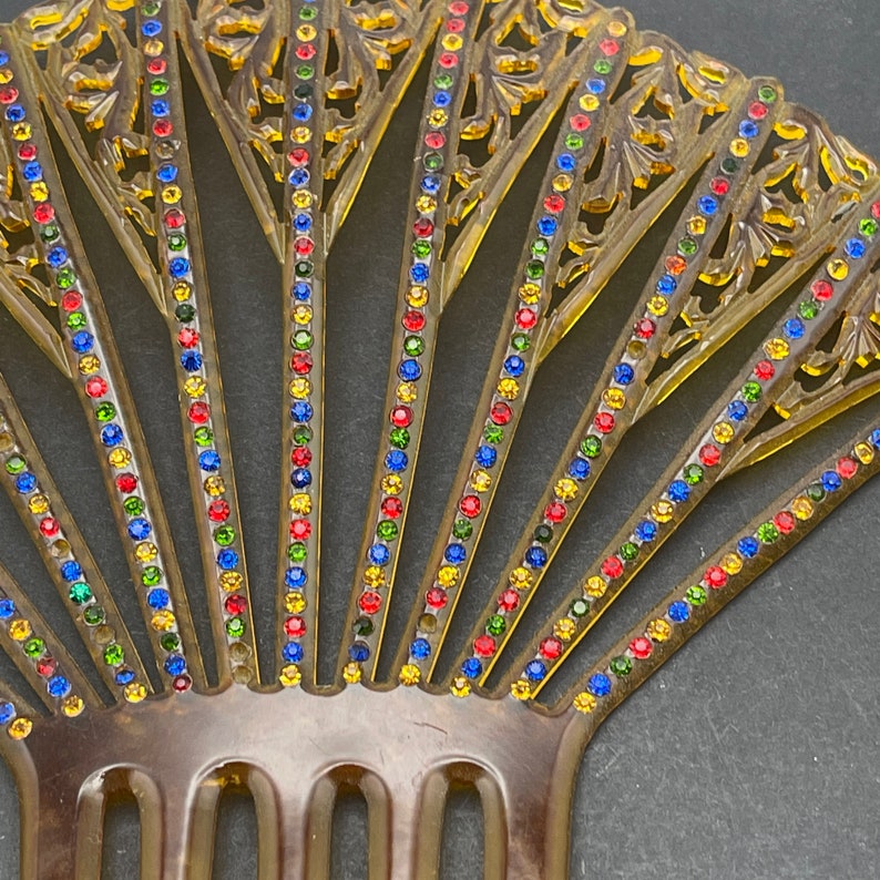 May include: A brown plastic hair comb with a fan-shaped design. The comb is decorated with rows of colorful rhinestones in red, yellow, blue, and green.