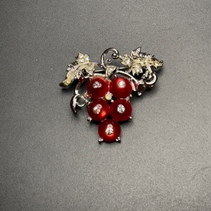 May include: A silver-toned brooch in the shape of a grape cluster. The brooch features five red, round, gem-like grapes with small, clear crystals. The leaves and vines are also silver-toned with clear crystal accents.