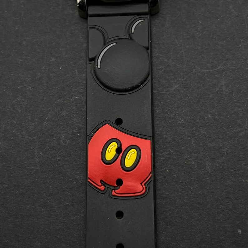 May include: Black silicone watch band with a red and yellow Mickey Mouse design. The band features a raised Mickey Mouse head and a raised Mickey Mouse in his signature red shorts.