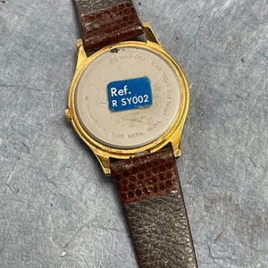 Pu&ograve; includere: Orologio da polso dorato con cinturino in pelle marrone. Il retro dell'orologio ha una lunetta in acciaio inossidabile con il testo "Ref. R SY002" stampato su un adesivo blu.