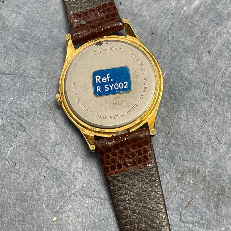Pu&ograve; includere: Orologio da polso dorato con cinturino in pelle marrone. Il retro dell'orologio ha una lunetta in acciaio inossidabile con il testo "Ref. R SY002" stampato su un adesivo blu.