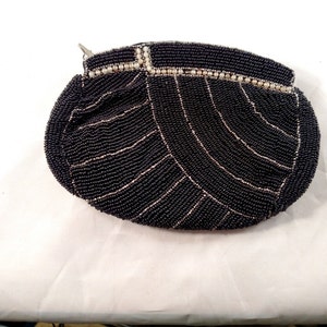 Puede incluir: Un bolso de mano negro con cuentas y cremallera plateada. El bolso está cubierto de pequeñas cuentas negras con líneas de acento plateadas. El borde superior está decorado con pequeñas cuentas blancas y un cierre plateado.