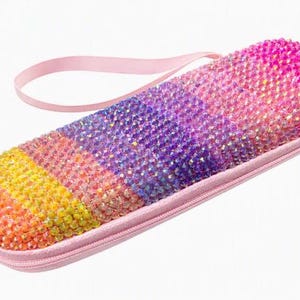May include: A rectangular zippered case featuring a vibrant gradient of multi-colored crystals in shades of orange, yellow, pink, and purple. The exterior is fully encrusted with reflective stones, and the case includes a matching light pink wrist strap.