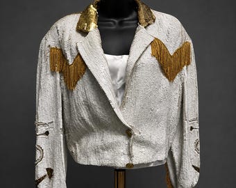 May include: A white sequined jacket featuring a gold sequined collar, gold fringe detailing across the chest, and long sleeves. The jacket has a single button closure at the front and is displayed on a mannequin, showcasing a white inner lining.