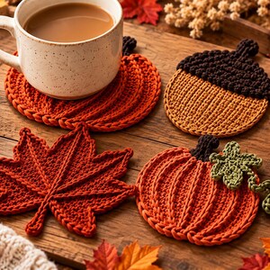 Könnte beinhalten: Ein Set aus drei gehäkelten Untersetzern mit herbstlichen Motiven: ein leuchtend orangefarbener Kürbis, eine braune Eichel mit strukturierter Kappe und ein dunkelrotes Ahornblatt. Jeder Untersetzer ist detailliert gestrickt und zeigt klare Formen.