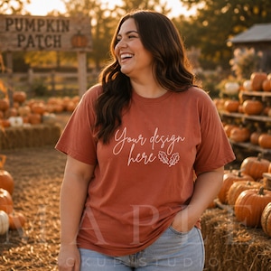 May include: A rust-colored short-sleeve t-shirt featuring the white cursive text Your design here followed by a small outline of two oak leaves. The shirt is worn with light blue denim jeans, creating a casual look suitable for autumn.