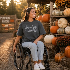 May include: A charcoal gray short-sleeve t-shirt with the white text Your design here and a small leaf graphic. The shirt is worn with light blue denim jeans while seated in a black wheelchair, positioned in an outdoor pumpkin patch setting.