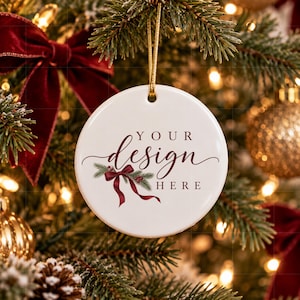 May include: A round white ceramic ornament hanging from a gold string. The ornament features the text "YOUR design HERE" in dark red script and serif fonts, accompanied by a graphic illustration of green pine needles and a dark red ribbon bow.