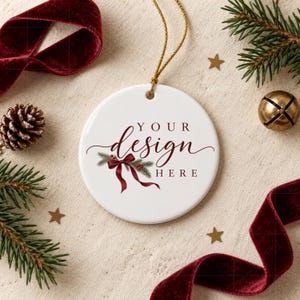 Könnte beinhalten: Ein runder, weißer Keramik-Weihnachtsanhänger mit goldener Aufhängeschnur und dem aufgedruckten Text "YOUR DESIGN HERE" in dunkelroter Schrift, verziert mit einer Grafik eines grünen Tannenzweigs, der mit einer dunkelroten Schleife gebunden ist.
