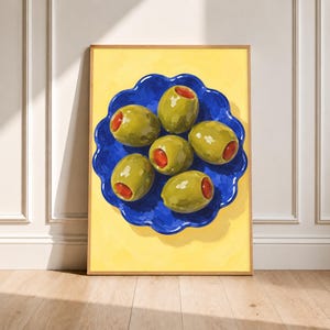 Può includere: Una stampa d'arte incorniciata che raffigura un dipinto vivace di sei olive verdi ripiene di peperone disposte su un piatto blu intenso con bordi smerlati. Il piatto è posizionato su uno sfondo giallo brillante, creando un contrasto cromatico netto.