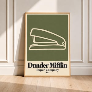 May include: A rectangular wall art print featuring a dark olive green background with a cream-colored line drawing of a stapler. Below the graphic, the text reads Dunder Mifflin Paper Company Inc. in bold black serif lettering on a cream base.