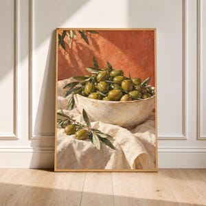 Op de afbeelding: Een ingelijste wandkunstprint met een rustieke, lichtgekleurde keramische kom gevuld met groene olijven en olijftakken met bladeren. De kom staat op een gedrapeerde, textuurrijke witte doek tegen een warme, terracotta-kleurige muur met schaduwen.