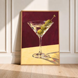 May include: A framed art print featuring a martini glass filled with a clear liquid and three green olives on a gold-coloured cocktail pick. The glass sits on a light yellow surface against a deep burgundy background with visible brushstrokes.