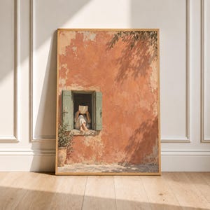 May include: A framed wall art print depicting a person sitting in a window frame of a weathered terracotta-colored building, reading an open book. The scene features sage green wooden shutters, a potted plant, and shadows of tree branches.