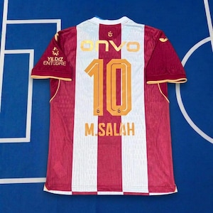 Puede incluir: Una camiseta deportiva de manga corta con rayas verticales granates y blancas. La parte posterior muestra el texto dorado "ONVO" sobre el número "10" y el nombre "M. SALAH" en dorado. Un logotipo "YILDIZ ENTEGRE" está impreso en la manga derecha.