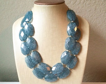 May include: A chunky blue statement necklace displayed on a mannequin. The necklace features large, faceted, irregular-shaped beads strung together with silver-toned connectors. The beads have a marbled appearance, and the necklace is a bold accessory.
