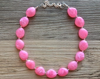 May include: A pink statement necklace featuring large, oval-shaped beads with a marbled effect. The beads are strung together with small silver-colored connectors and a chain clasp. The necklace is displayed on a wooden surface.