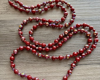 May include: A long red beaded necklace with iridescent accents. The necklace is made of round beads and is strung on a silver chain.