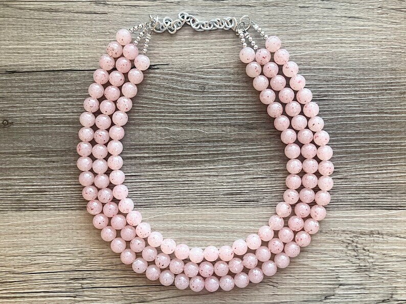 May include: A multi-strand necklace made of pink beads with a speckled pattern. The necklace is made of three strands of beads and has a silver clasp.
