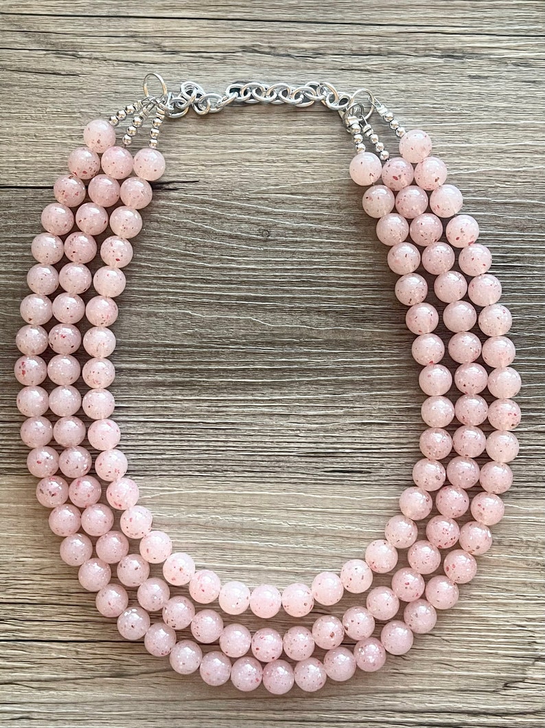 May include: A three-strand pink beaded necklace with a silver chain clasp. The beads are round and have a speckled texture.