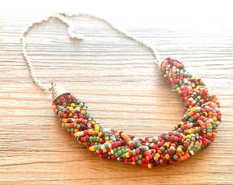 May include: A colorful beaded necklace with a silver chain. The necklace features a thick, braided design of small, round beads in various colors, including red, orange, yellow, green, and black. The necklace is displayed on a light brown wooden surface.