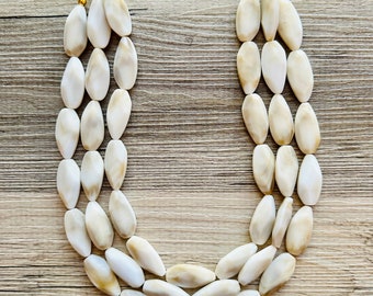 May include: A three-strand statement necklace featuring large, oval-shaped off-white beads. The beads are strung on a gold-tone chain and clasps. The necklace is displayed on a light wood-grain surface.