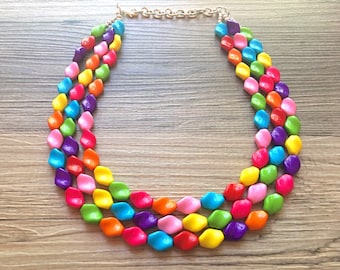 Puede incluir: Un collar de múltiples hilos hecho de cuentas de plástico facetadas y de colores. El collar está hecho de tres hilos de cuentas, cada hilo es de un color diferente. Las cuentas están dispuestas en un patrón repetitivo de rosa, amarillo, verde, morado, naranja y azul.