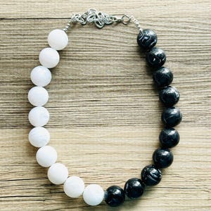 May include: A necklace featuring a split design with smooth white beads on one side and black marbled beads on the other. The beads are connected by a silver-toned metal chain and clasp mechanism at the top, creating a circular arrangement.