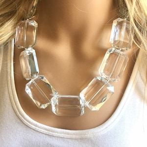 May include: A clear, chunky necklace featuring large, rectangular crystal-like beads. The beads are strung together, creating a statement piece. The necklace is worn against a white tank top, highlighting the jewelry's sparkle and design.