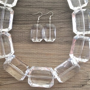 May include: A clear, faceted necklace and matching earrings set. The necklace features large, rectangular beads strung together. The earrings are rectangular with silver hooks. The jewelry is displayed on a wooden surface.