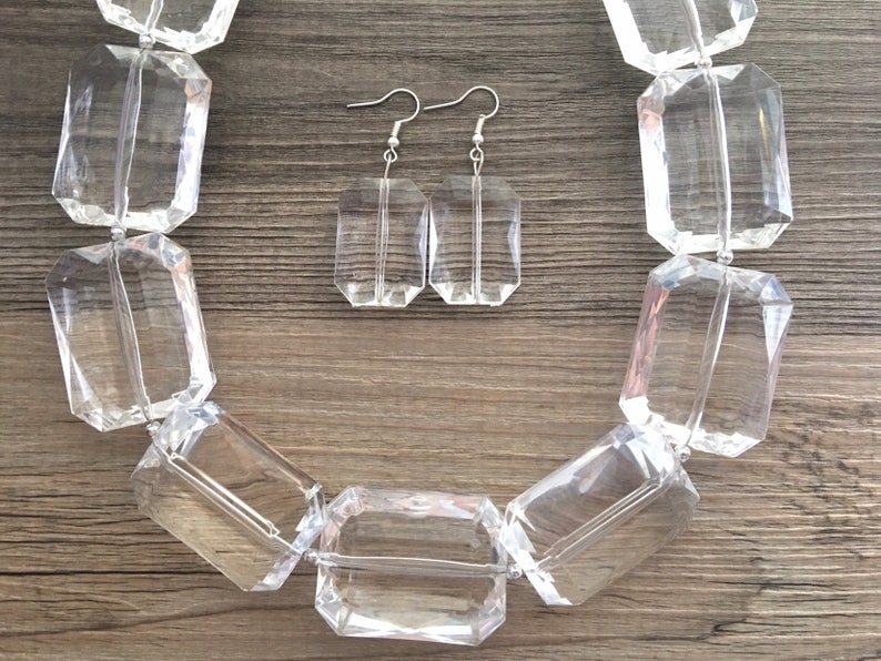 May include: A clear, faceted necklace and matching earrings set. The necklace features large, rectangular beads strung together. The earrings are rectangular with silver hooks. The jewelry is displayed on a wooden surface.