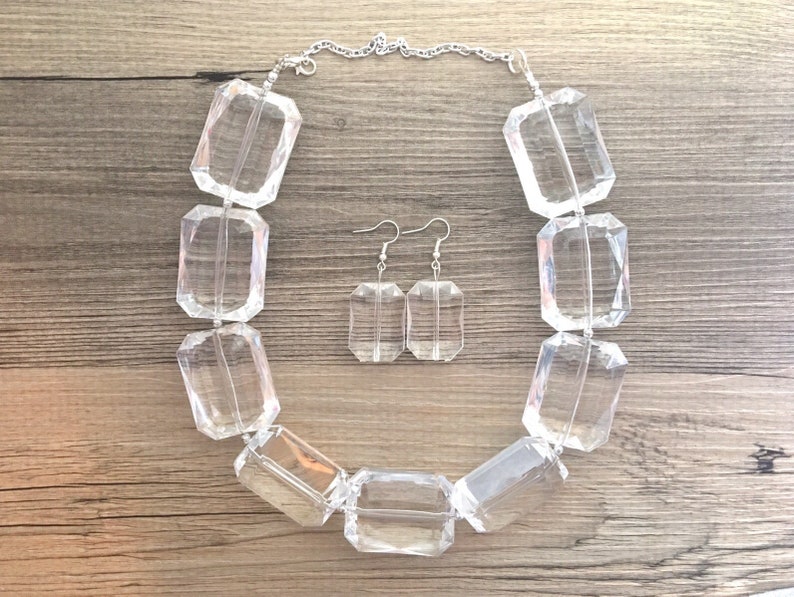 May include: A clear, rectangular bead necklace and matching earrings set. The necklace features large, faceted beads strung on a silver chain. The earrings have a similar design, with rectangular clear beads suspended from silver hooks.