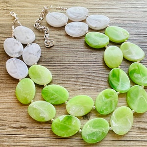 May include: A statement necklace featuring large, oval-shaped beads in shades of white and green. The beads are strung on a silver-tone chain with a lobster clasp closure. The necklace is displayed on a wooden surface.