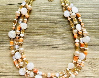 May include: A multi-strand necklace with gold, white, and peach colored beads. The necklace is made of various sizes and shapes of beads, including round, oval, and flat.