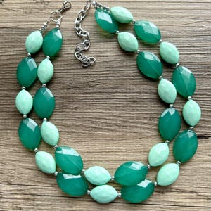 May include: A double-strand necklace with oval-shaped beads in shades of green. The necklace is made of light green and dark green beads.