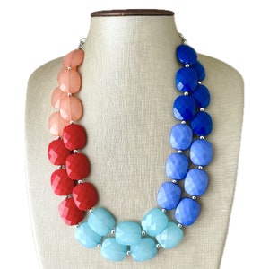 May include: A multi-strand necklace with large, faceted, square-shaped beads in shades of peach, red, blue, and turquoise. The necklace is made of plastic and has a silver-toned metal clasp.