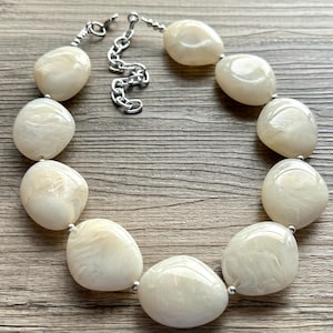 May include: A cream-colored statement necklace featuring large, oval-shaped beads with a marbled effect. The beads are strung together with silver-toned metal findings and a chain for adjustable length. The necklace is displayed on a wooden surface.